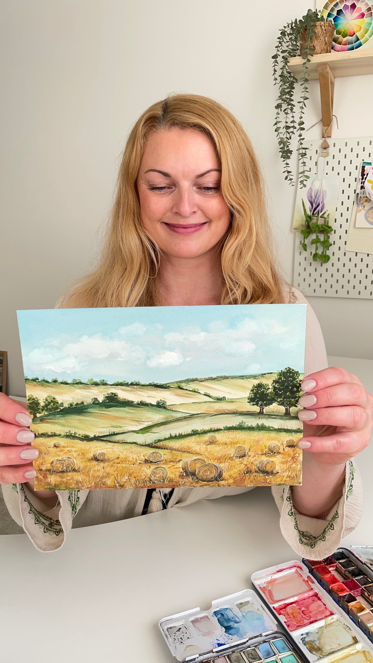Žena držící originální akvarelovou malbu letního zlatého pole s balíky slámy, sedící u stolu s paletou akvarelových barev.