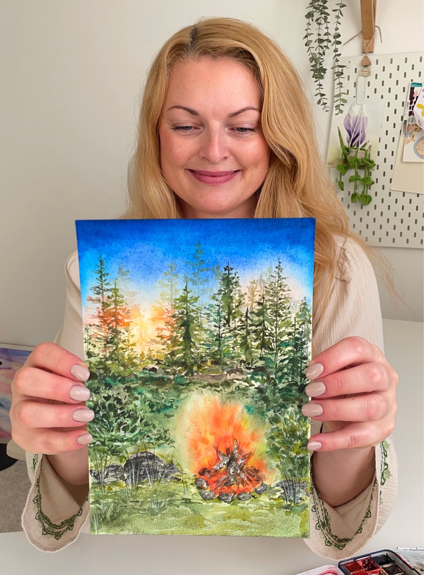 Žena držící originální akvarelovou malbu lesního táboráku při západu slunce, usmívající se u pracovního stolu.