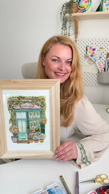 Žena usmívající se do kamery a držící zarámovanou akvarelovou ilustraci květinářství v přírodním dřevěném rámu, sedící u pracovního stolu s malířskými pomůckami.