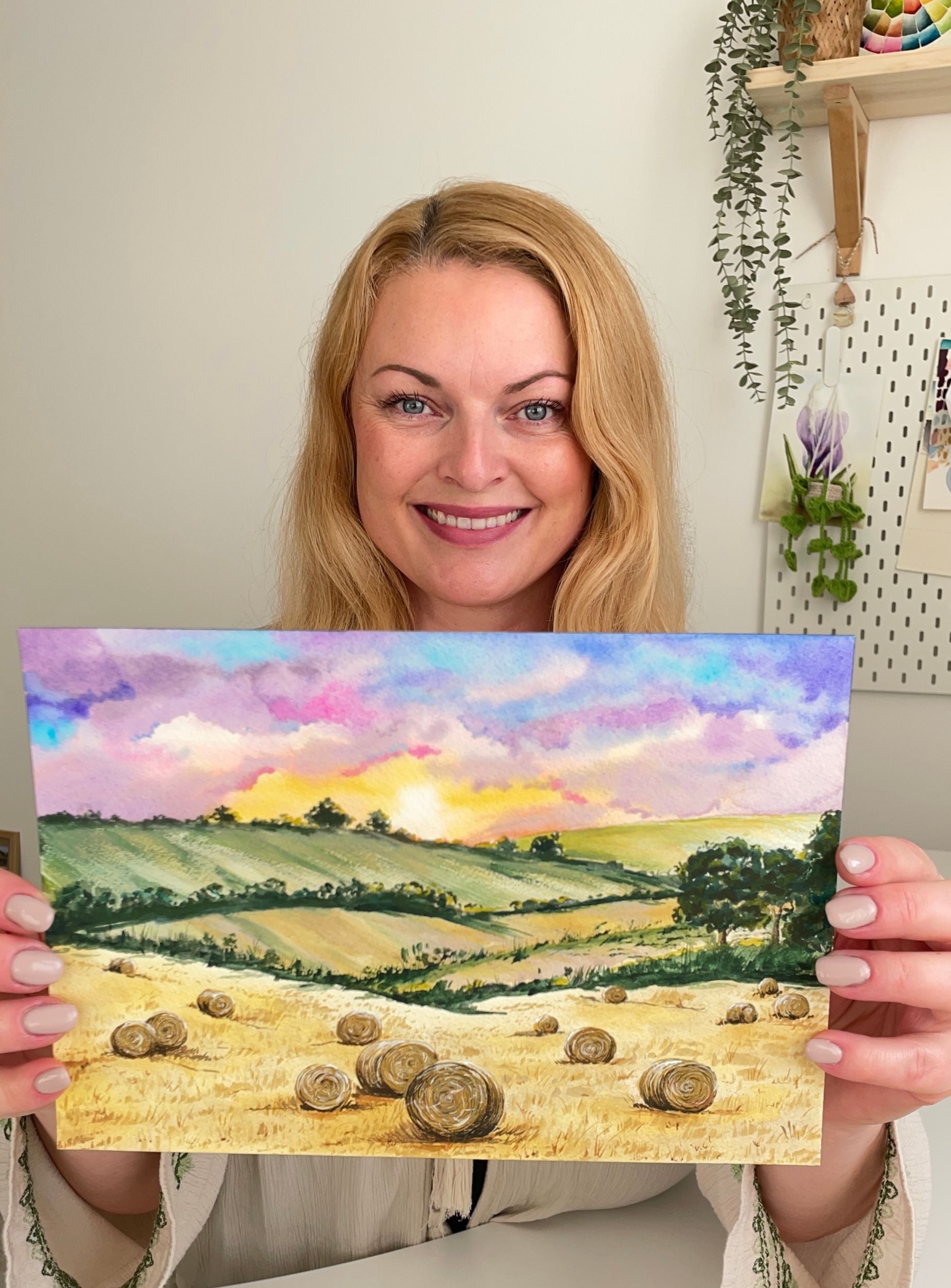 Žena usmívající se do kamery a držící originální akvarelovou malbu letního zlatého pole s balíky slámy.