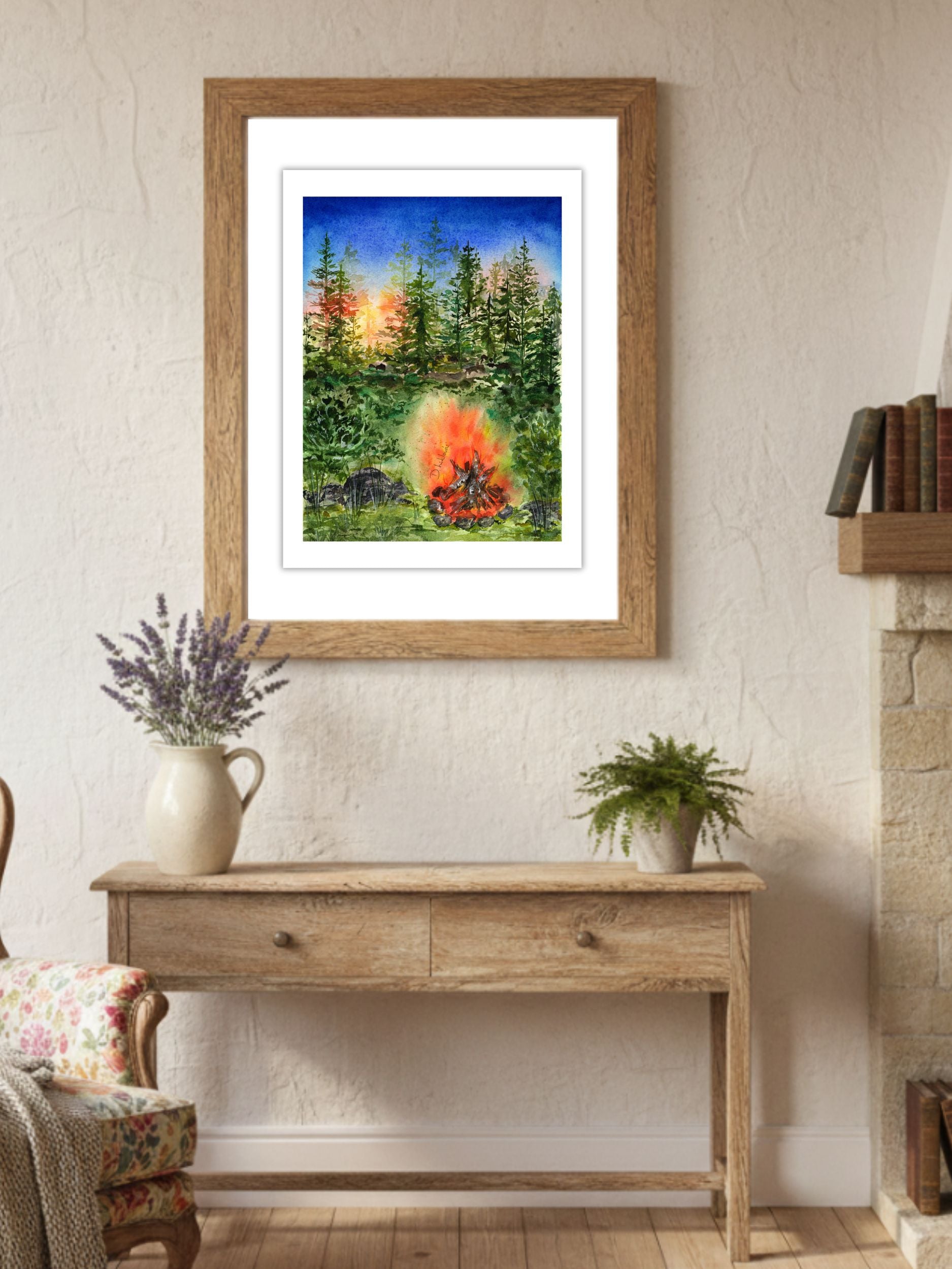 Akvarelový obraz lesního ohně zavěšený v dřevěném rámu v útulném venkovském interiéru. Fine art tisk s motivem lesa při západu slunce a praskajícího ohně, vystavený nad dřevěným stolem s levandulí a zelenou rostlinou.