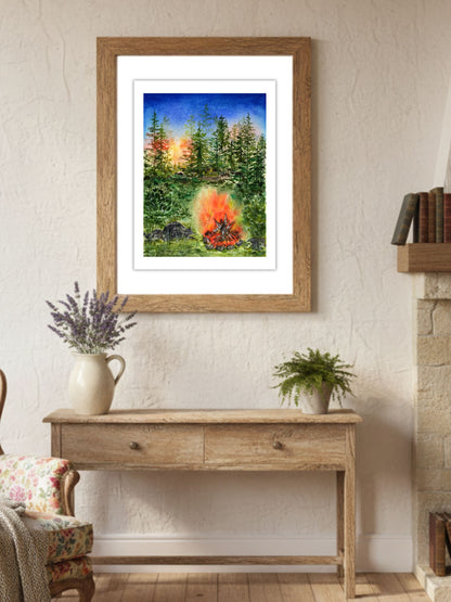 Akvarelový obraz lesního ohně zavěšený v dřevěném rámu v útulném venkovském interiéru. Fine art tisk s motivem lesa při západu slunce a praskajícího ohně, vystavený nad dřevěným stolem s levandulí a zelenou rostlinou.