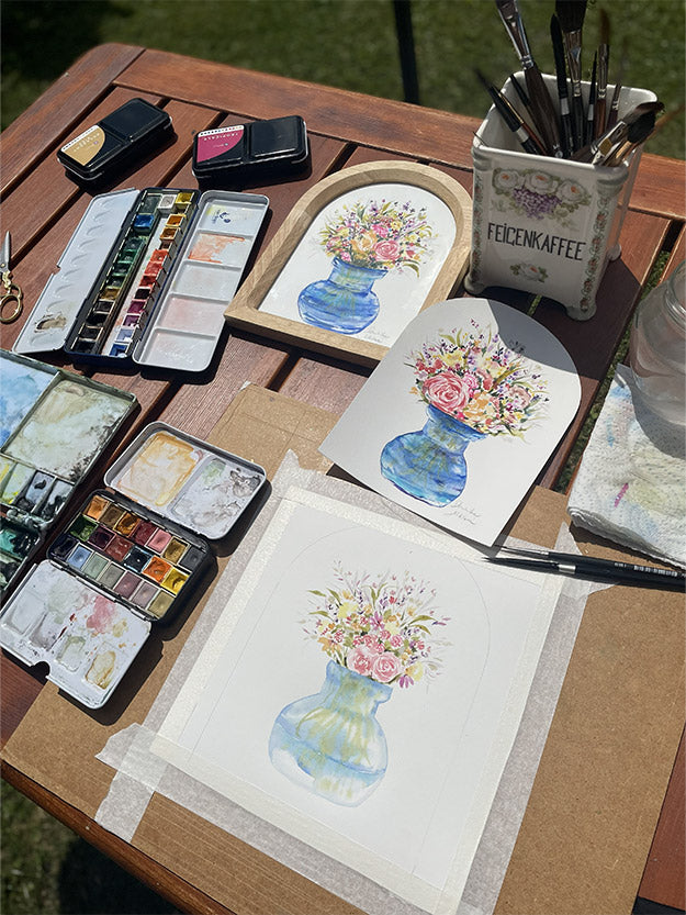Tvoření akvarelové malby květin ve váze na venkovním stole, s paletami, štětci a rozpracovanými ilustracemi.